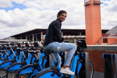 Siyah gömlekli ve yırtık pantolonlu Hintli bir adam bisiklet paylaşım istasyonunda oturmuş gülümsüyor..