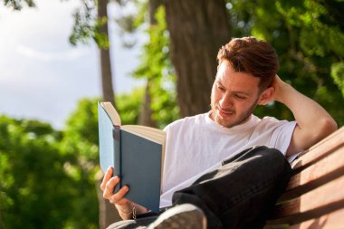 Bir adam bankta oturmuş kitap okuyor. Kitap mavi ve adam beyaz bir gömlek giyiyor. Sahne huzurlu ve rahatlatıcı, adam ağaçların gölgesinde kitap okumaktan zevk alıyor.