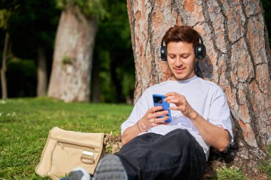 Latin bir adam parkta çimenlerde oturur, ağaca yaslanır. Rahatlıyor ve gevşiyor, akıllı telefonunu günlük stresten kurtulmak için kullanıyor..