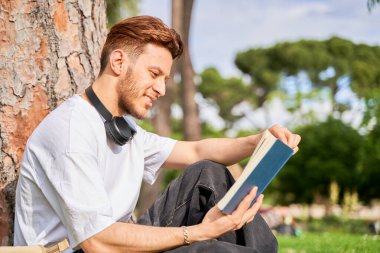 Bir adam bir ağacın altında oturmuş kitap okuyor. Kulaklık takıyor ve yüzünde bir gülümseme var.