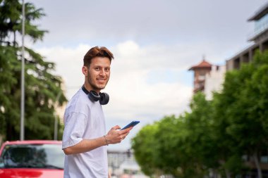 Bir adam yol kenarında duruyor, telefon elinde, yola bakıyor, şehirde bir taksi ya da otobüs bekliyor, teknolojiyi kentsel hareketlilikle harmanlıyor..