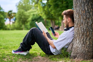 Bir adam bir ağacın altında oturuyor ve cep telefonu tutarken kitap okuyor. Rahatlama ve rahatlama kavramı, adam dışarıda vakit geçirmekten ve hem kitap okumaktan zevk alıyor.