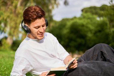 Genç bir Latin adam parkta çimenlerde oturur, kulaklıklarıyla müzik dinlerken bir kitaba dalmış, sakin bir açık hava ortamında rahatlık ve keyif katar..
