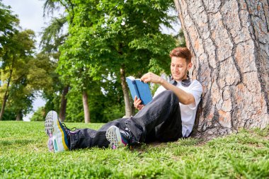 Bir adam parkta çimenlerde oturur, bir ağaca yaslanır kitap okurken, doğada huzurlu bir anın tadını çıkarır..