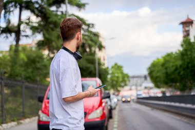 Bir adam yol kenarında duruyor, telefon elinde, yola bakıyor, şehirde bir taksi ya da otobüs bekliyor, teknolojiyi kentsel hareketlilikle harmanlıyor..