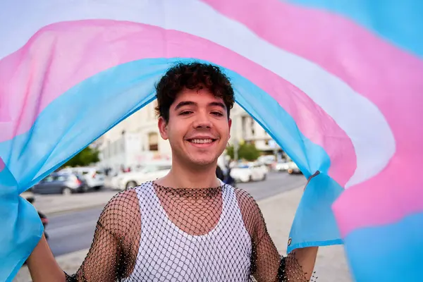 Bir adam gülümsüyor ve elinde bir trans bayrak tutuyor. Arkasında bayrak var ve beyaz bir gömlek giyiyor.
