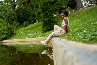 Parkta genç bir Afrikalı kadının portreleri, arka planda dingin bir gölle duruyor, doğrudan kameraya bakıyor, güven ve sükunet yayıyor..