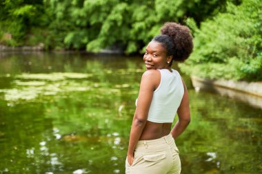 Parkta genç bir Afrikalı kadının portreleri, arka planda dingin bir gölle duruyor, doğrudan kameraya bakıyor, güven ve sükunet yayıyor..