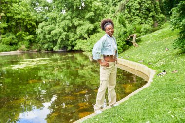 Parkta genç bir Afrikalı kadının portreleri, arka planda dingin bir gölle duruyor, doğrudan kameraya bakıyor, güven ve sükunet yayıyor..