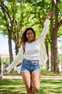 Kaygısız Latin kız parkta gülüp dans ediyor güneşli yaz gününün tadını çıkarıyor, el kaldırıyor.