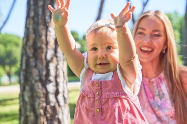 Güneşli bir parkta elleri havada gülümseyen bebek, arka planda annesiyle neşeli bir anın tadını çıkarıyor.