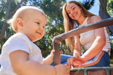 Bir kadın kucağında bebekle bankta oturuyor. Bebek gülümsüyor ve kadın da ona gülümsüyor. Sahne mutlu ve neşeli.