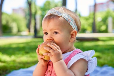 Parkta battaniyede oturan, elmayı tutan ve ısıran bir bebek. Bebeğin kafasında saç bandı ve arkasında yeşil ağaçlar olan pembe bir elbise var..