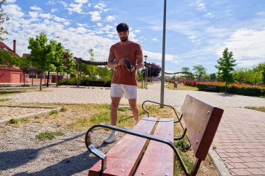 Sakallı genç bir sporcu antrenmanına başlamadan önce oturmak ve hazırlanmak için bir banka doğru yürür. Açık hava ortamındaki bir odak ve hazırlık anını somutlaştırır..