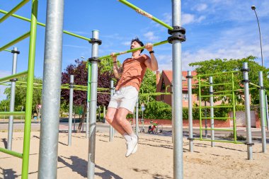 Genç bir atlet, açık hava idman ortamında güç ve adanmışlığını göstermek için açık hava spor parkında barfiks çekiyor..