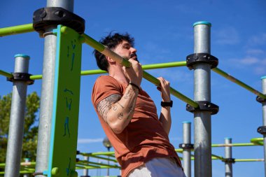 Genç bir atlet, açık hava idman ortamında güç ve adanmışlığını göstermek için açık hava spor parkında barfiks çekiyor..