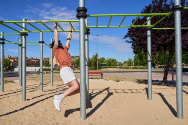 Genç bir atlet, açık hava idman ortamında güç ve adanmışlığını göstermek için açık hava spor parkında barfiks çekiyor..