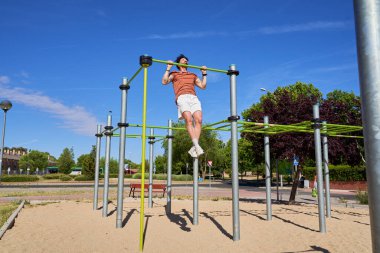 Genç bir atlet, açık hava idman ortamında güç ve adanmışlığını göstermek için açık hava spor parkında barfiks çekiyor..