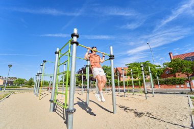 Genç bir atlet, açık hava idman ortamında güç ve adanmışlığını göstermek için açık hava spor parkında barfiks çekiyor..