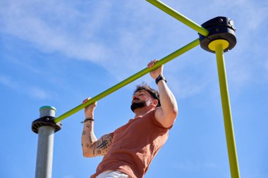 Genç bir atlet, açık hava idman ortamında güç ve adanmışlığını göstermek için açık hava spor parkında barfiks çekiyor..