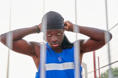 Mavi askılı bir adam siyah bir bandana takıyor. Bir çitin önünde duruyor.