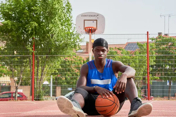 Mavi gömlekli bir adam basketbol topu tutuyor. Gülümsüyor ve basketbol potasının önünde duruyor.