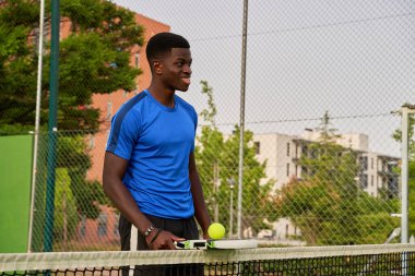 Tenis kortunda elinde tenis raketi ve tenis topu tutan bir adam var..