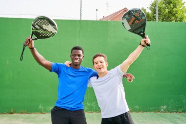 İki adam raket tutuyor ve gülümsüyor. Tenis kortunda poz veriyorlar.