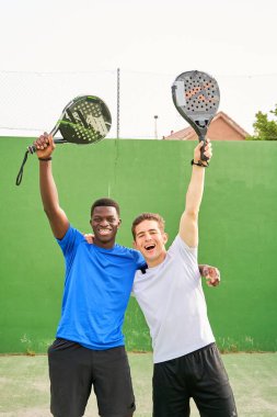 İki adam raket tutuyor ve gülümsüyor. Tenis kortunda poz veriyorlar.