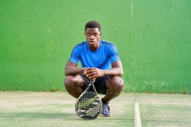 Mavi gömlekli bir adam elinde tenis raketiyle yerde oturuyordur. Kameraya ciddi bir ifadeyle bakıyor.