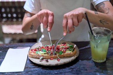 Bir insan çatal ve bıçakla sandviç yiyor. Sandviç tabakta ve yanında bir bardak limonata var.