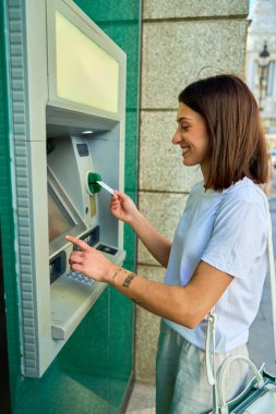 Şehir caddesinde ATM kullanan genç bir kadın yeşil bir el çantasıyla kartını yerleştirirken gülümsüyor..