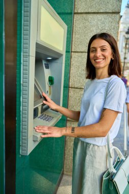 Şehir caddesinde ATM kullanan genç bir kadın. Elinde yeşil bir çantayla kameraya gülümsüyor..