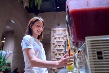 A woman is smiling as she fills a cup with orange juice from a dispenser. The scene is set in a restaurant or cafe, and the woman is enjoying her drink clipart