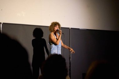 Kıvırcık saçlı beyaz kadın sanatçı, loş aydınlatılmış bir tiyatroda seyircilerle monolog yapıyor.