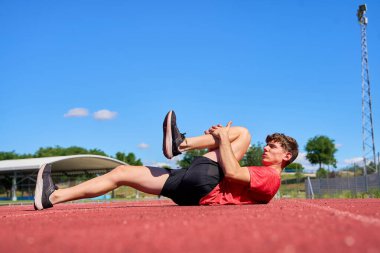 Bir adam bacağı bükülmüş ve ayağı dizinde yerde yatıyor. Kırmızı gömlek ve siyah şort giyiyor.