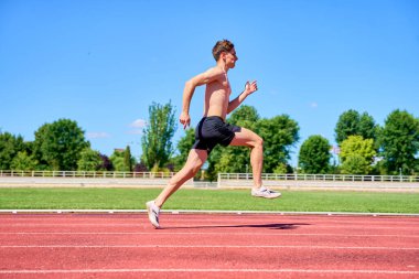 Bir adam kolları açık bir şekilde pistte koşuyor. Gökyüzü mavi ve çimenler yeşil