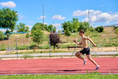 Arka planda mavi gökyüzü olan bir adam pistte koşuyor. Gökyüzü açık ve parlak, ve adam siyah bir gömlek ve şort giyiyor.
