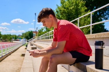 Teneffüste akıllı telefon kullanan bir sporcu tribünde oturuyor.