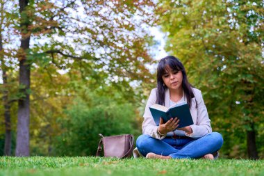 Latin bir kadın parkta çimlerin üzerinde oturmuş kitap okuyor.
