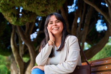 Gülümseyen Latin kadın parktaki ağaçların altında oturuyor.