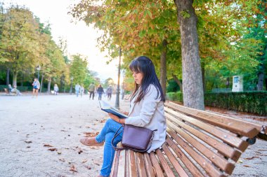 Latin bir kadın parkta bir bankta oturmuş kitap okuyor.