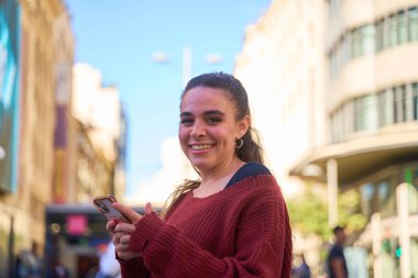 Güneşli bir şehir sokağında elinde akıllı telefonla gülümseyen beyaz bir kadın.