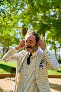 Güneşli bir parkta elinde kulaklıkla gülümseyen bir adam.