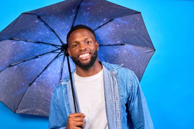 Üzerinde galaksi baskısı olan bir şemsiye tutan neşeli bir gencin, canlı mavi bir arkaplanda poz verirken çekilmiş bir portresi.