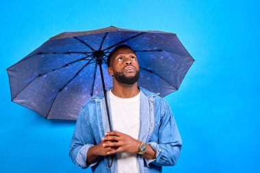 Genç Afrikalı Amerikalı bir adam elinde şemsiyeyle ayakta duruyor. Huzurlu bir mavi arka planda umutla yukarı bakıyorum. Dalgın dalgın dalgın dalgın dalgın dalgın. Yağmurun durmasını bekliyorum.