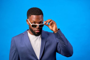 Çok zarif bir iş adamının portresi, parlak mavi bir zeminde poz verirken güneş gözlüğünü ayarlıyor.