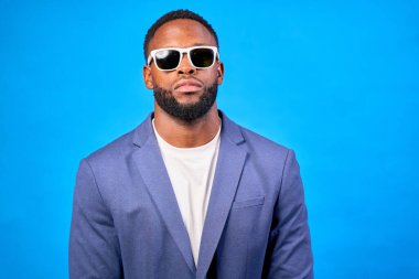 Güneş gözlüğü takan ve mavi takım elbiseli bir iş adamının stüdyo portresi canlı mavi bir arka plana karşı güven yayıyor.