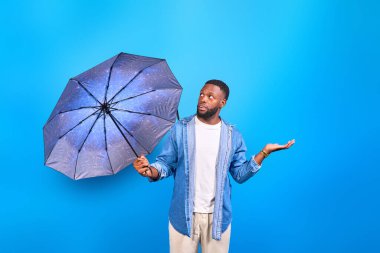 Şemsiye tutan bir gencin stüdyo fotoğrafı. Şaşırmış ve kafası karışmış görünüyor. Tek eliyle soru sorar gibi hareket ediyor.