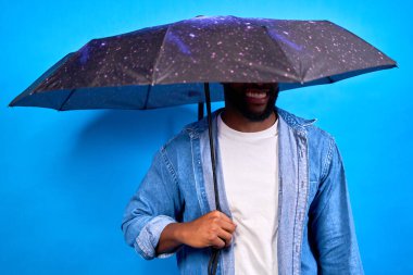 Uzay temalı bir şemsiye tutan gülümseyen bir adamın canlı mavi bir arka plana karşı stüdyo fotoğrafı.